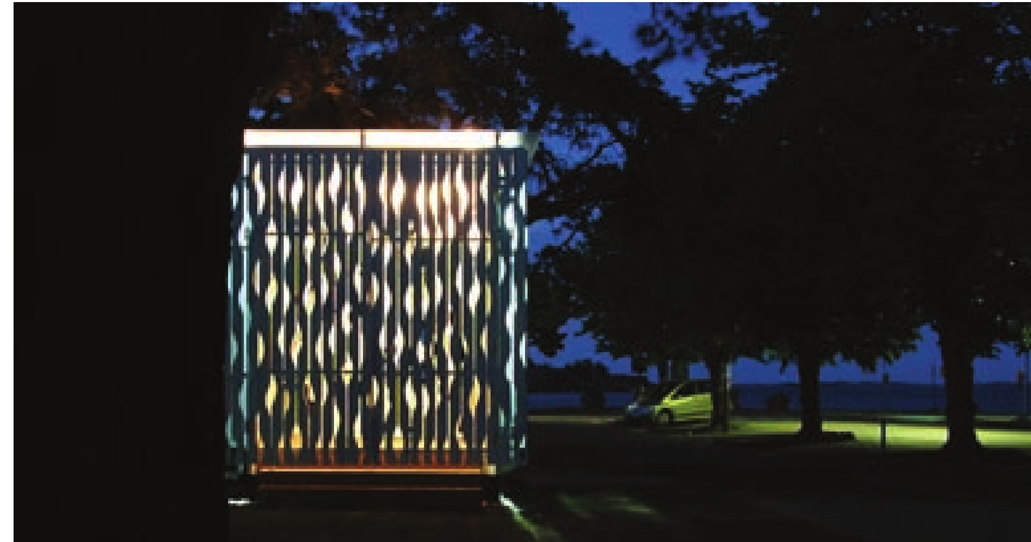 The finished kiosk at night, light spilling through the carved cladding so it reads as a lantern