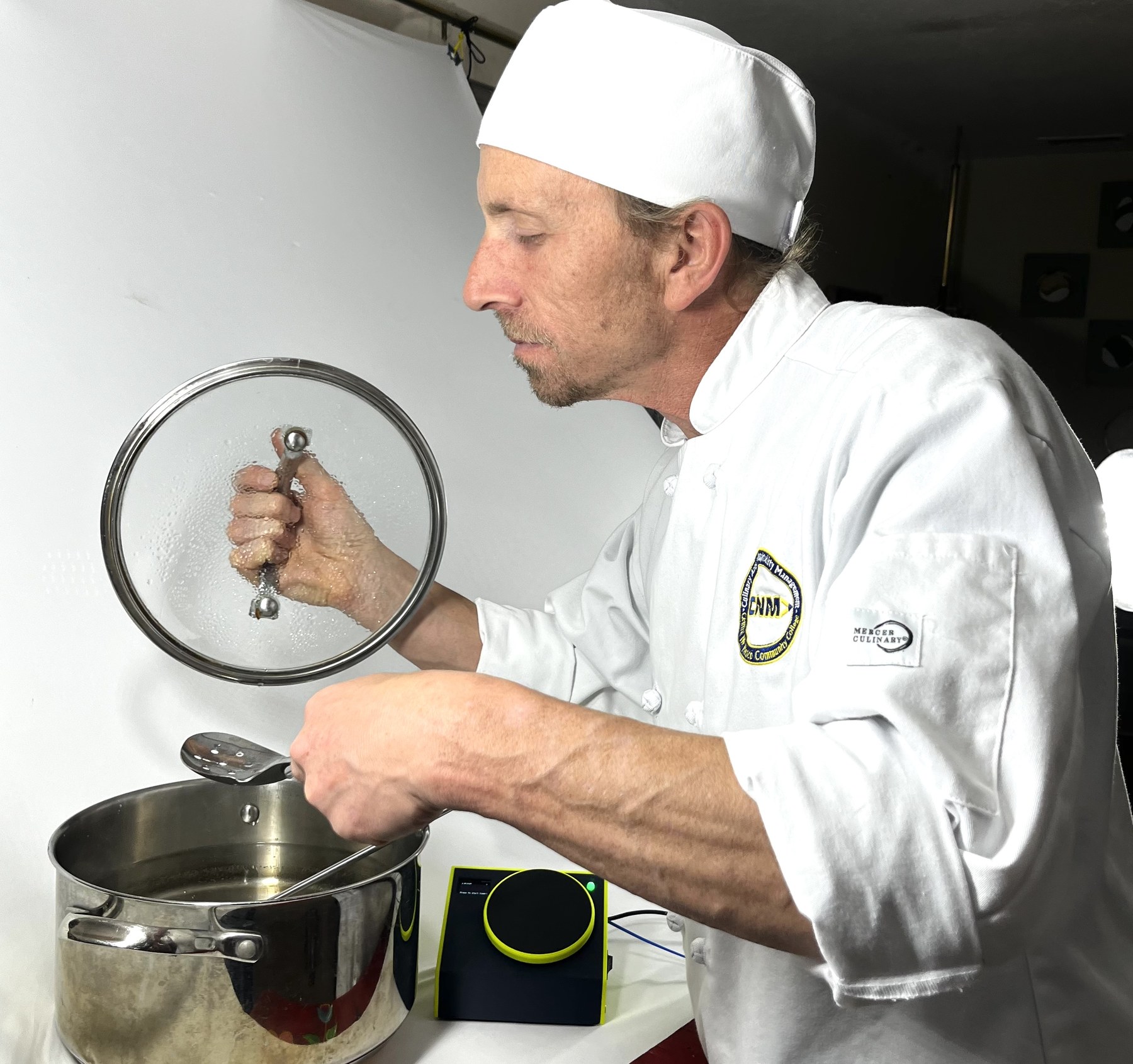 A cook in chef's whites lifting a glass lid to check the pot, the timer alongside
