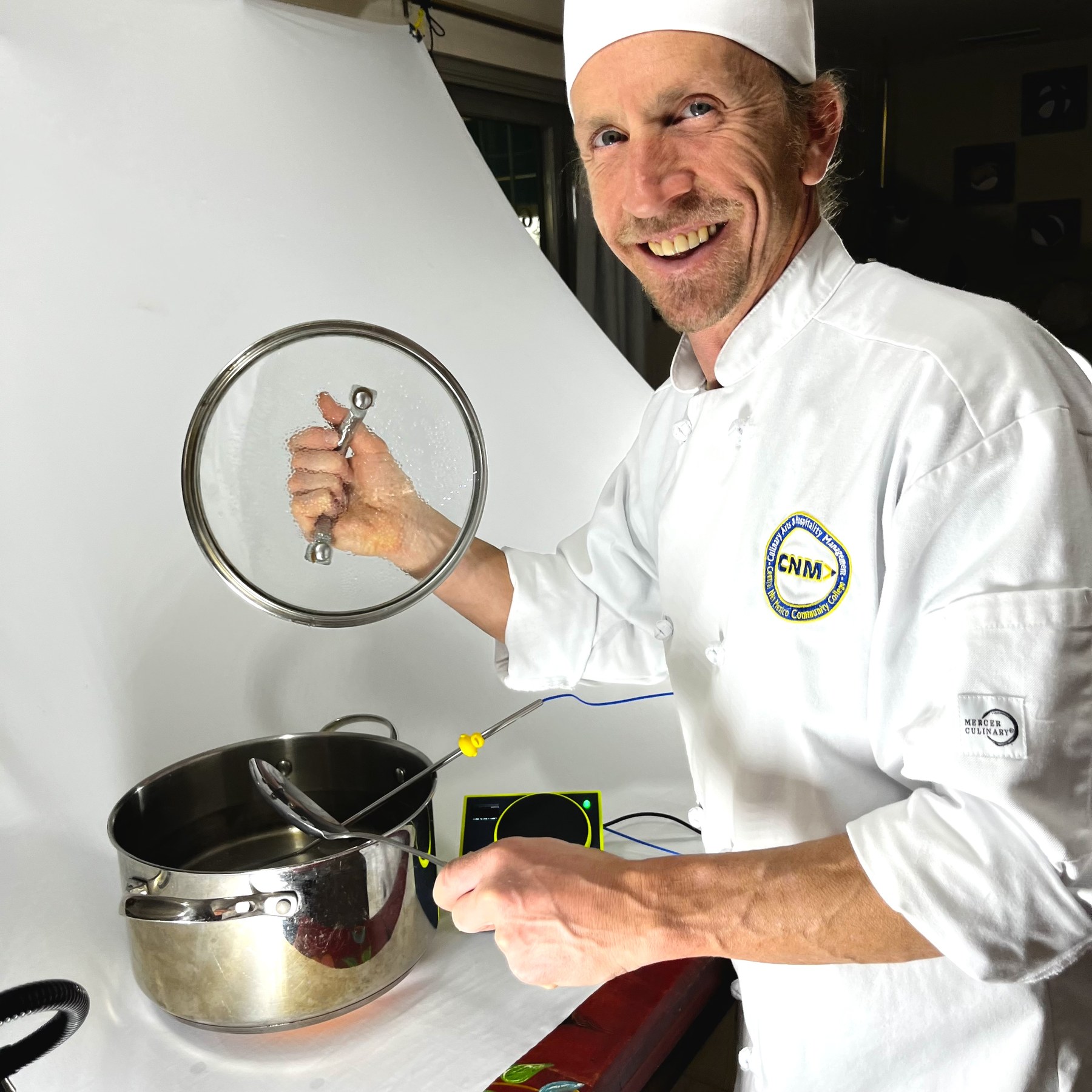 A smiling cook in a CNM Culinary Arts jacket working at the stove with the timer beside him