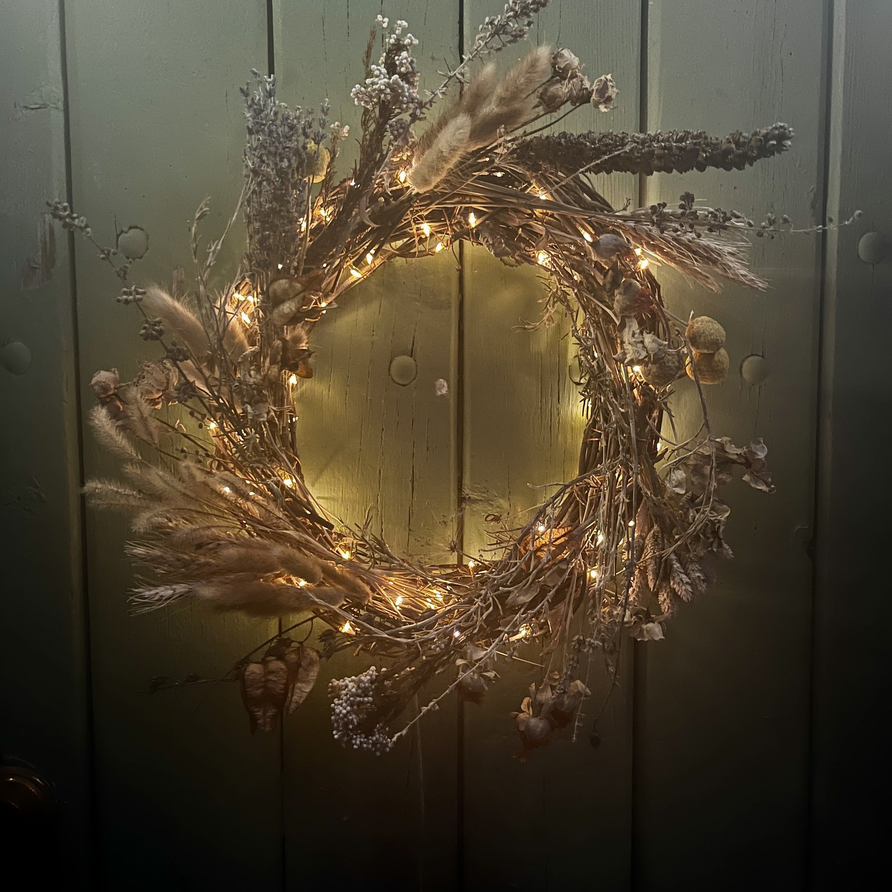 The finished wreath at night on a dark door, its fairy lights glowing warm through the dried grasses